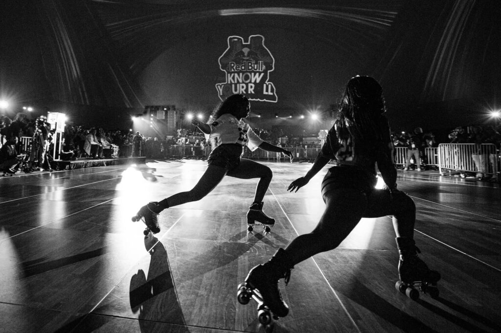 Two people roller skate on an indoor rink during a competition, with an audience and event branding visible in the background.