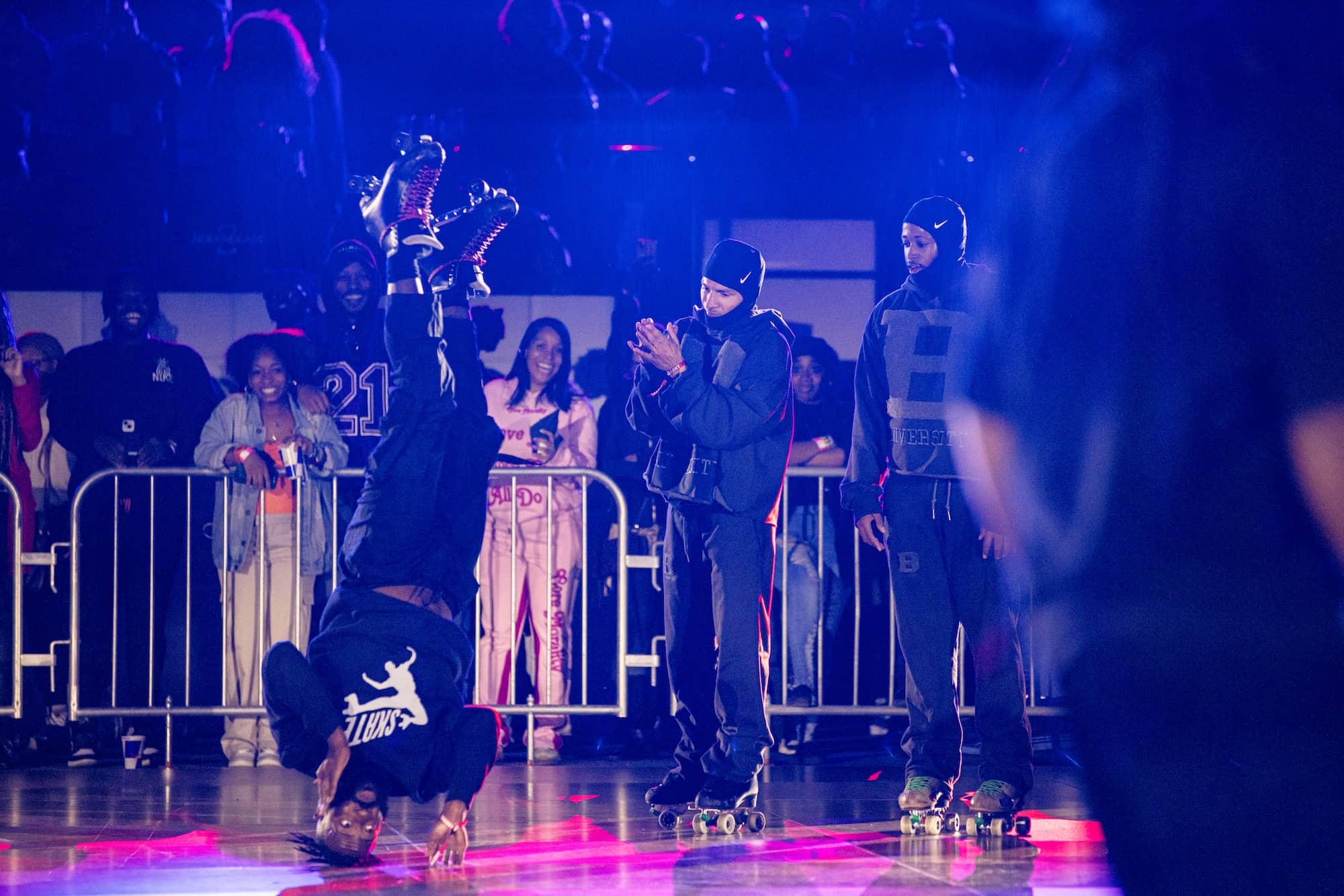 A person does a headstand on roller skates in front of a crowd at an indoor skating event, while others watch and clap.