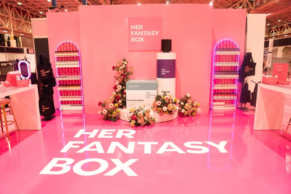 A pink exhibition booth with "HER FANTASY BOX" branding, oversized product props, floral decorations, and shelves of small boxes displayed on each side.