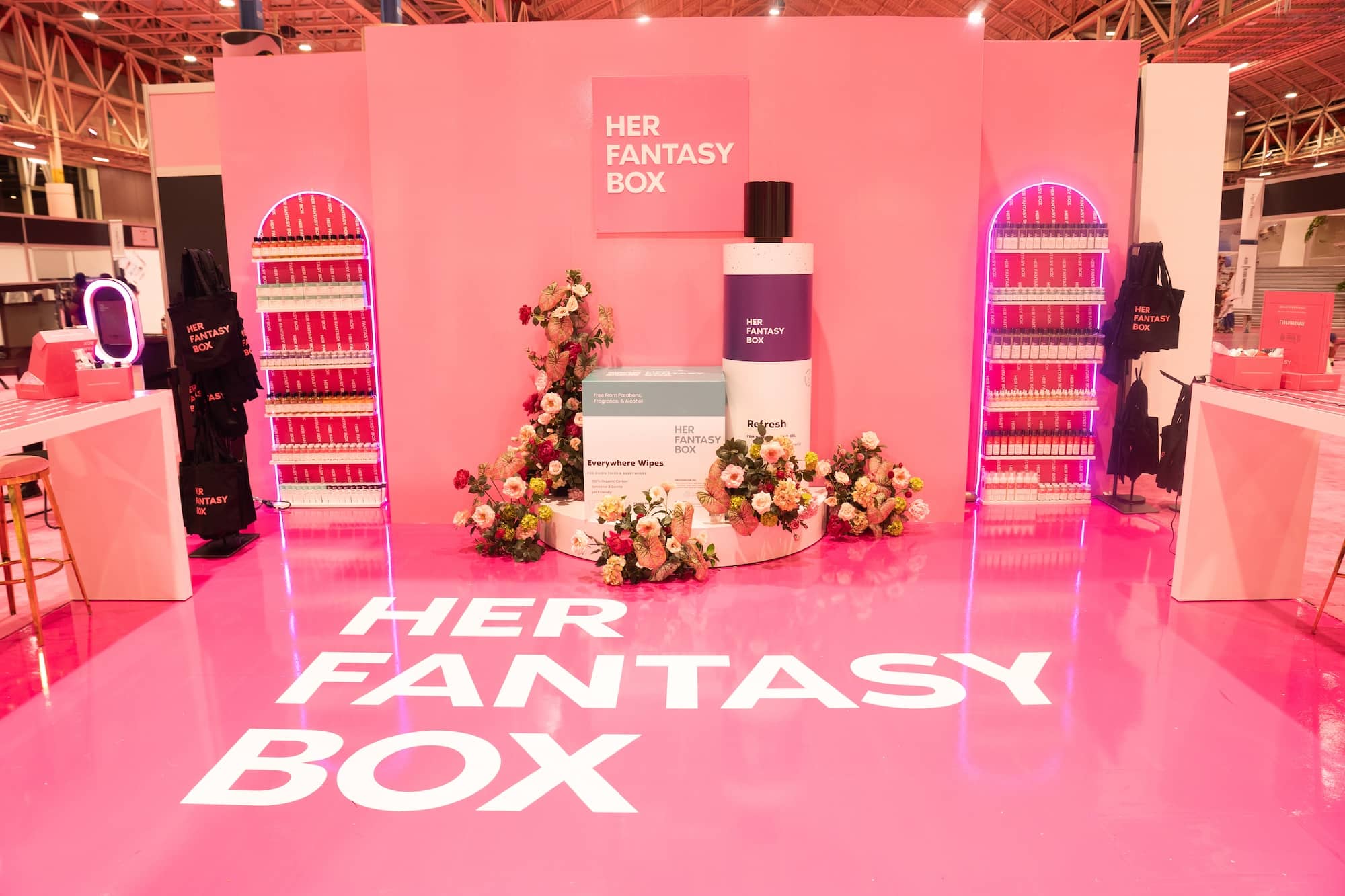 A pink exhibition booth with "HER FANTASY BOX" branding, oversized product props, floral decorations, and shelves of small boxes displayed on each side.