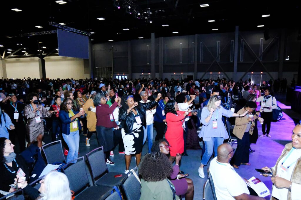 A large group of people stand and dance in a conference hall, with rows of chairs and a stage visible in the brightly lit room.