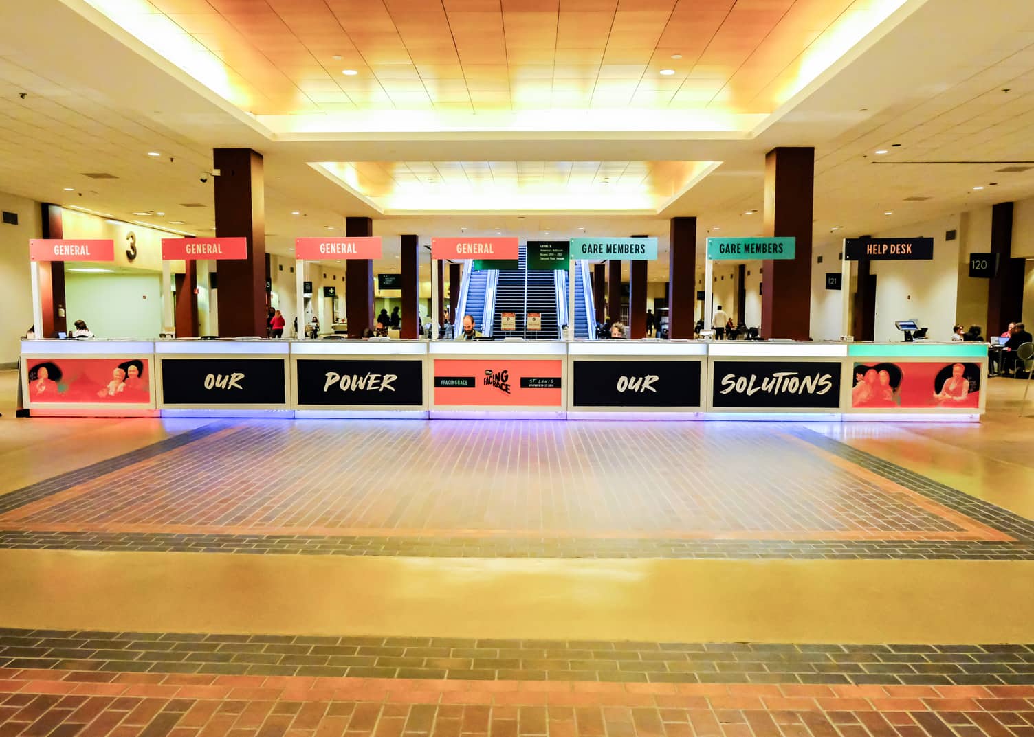 A brightly lit convention check-in area with labeled booths for General, GABE Members, and Help Desk; people sit behind the counters.