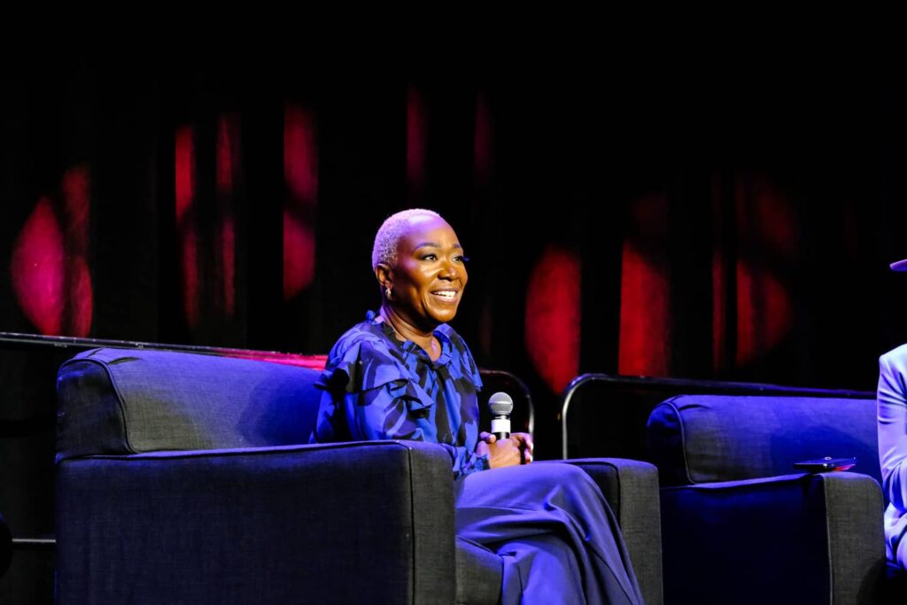 A person with short hair, wearing a blue outfit, sits on a dark armchair on stage holding a microphone, with a black and red background.