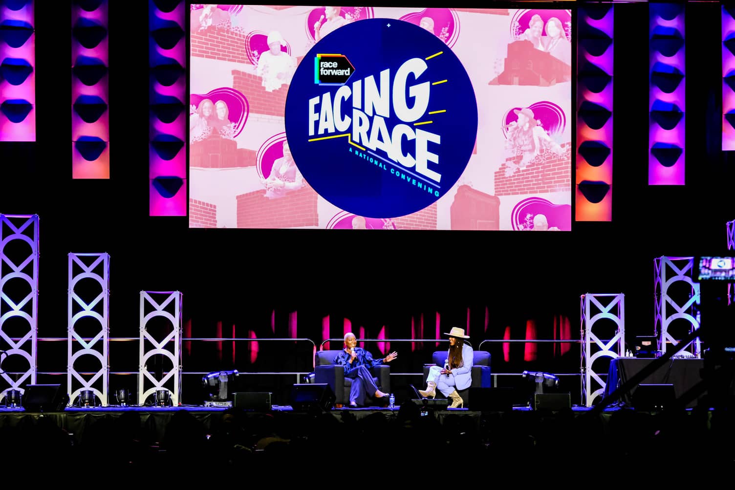 Two people are seated on stage in front of a large screen displaying "Facing Race" at a conference event with stage lighting and audience in the foreground.