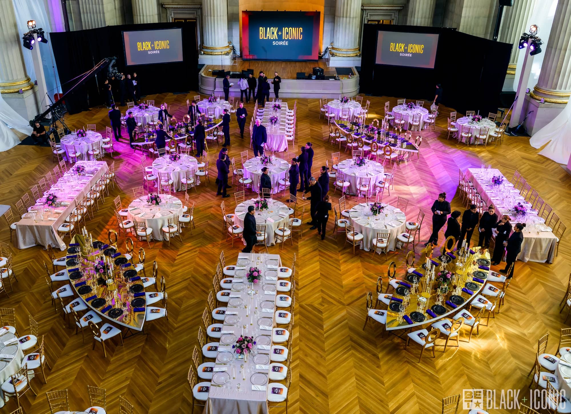People gather in an elegant hall set with round and rectangular tables for the Black Iconic Awards, with two large screens displaying the event name.