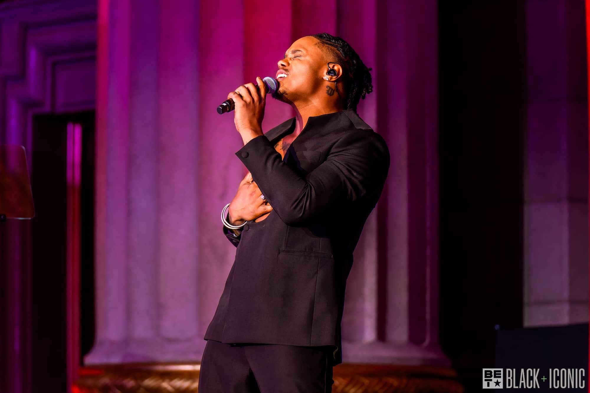 A man in a black suit sings passionately into a microphone on stage, with one hand on his chest and large columns in the background.