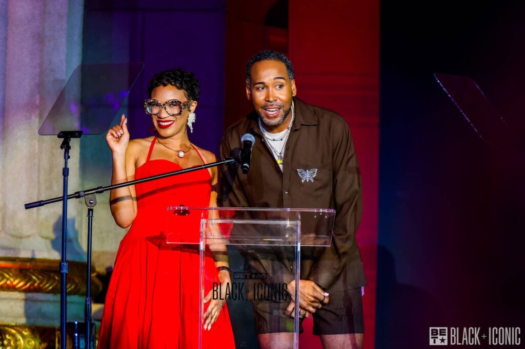 Two people stand at a clear podium on stage, one wearing a red dress and glasses, the other in a brown outfit, both smiling. The background features colorful lighting and BET Black + Iconic branding.