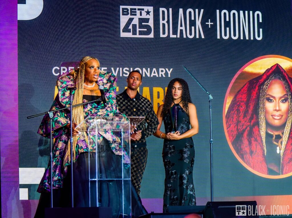 Three people stand on stage at a BET Black + Iconic event, with one person speaking at a podium and a large portrait image displayed on the screen behind them.