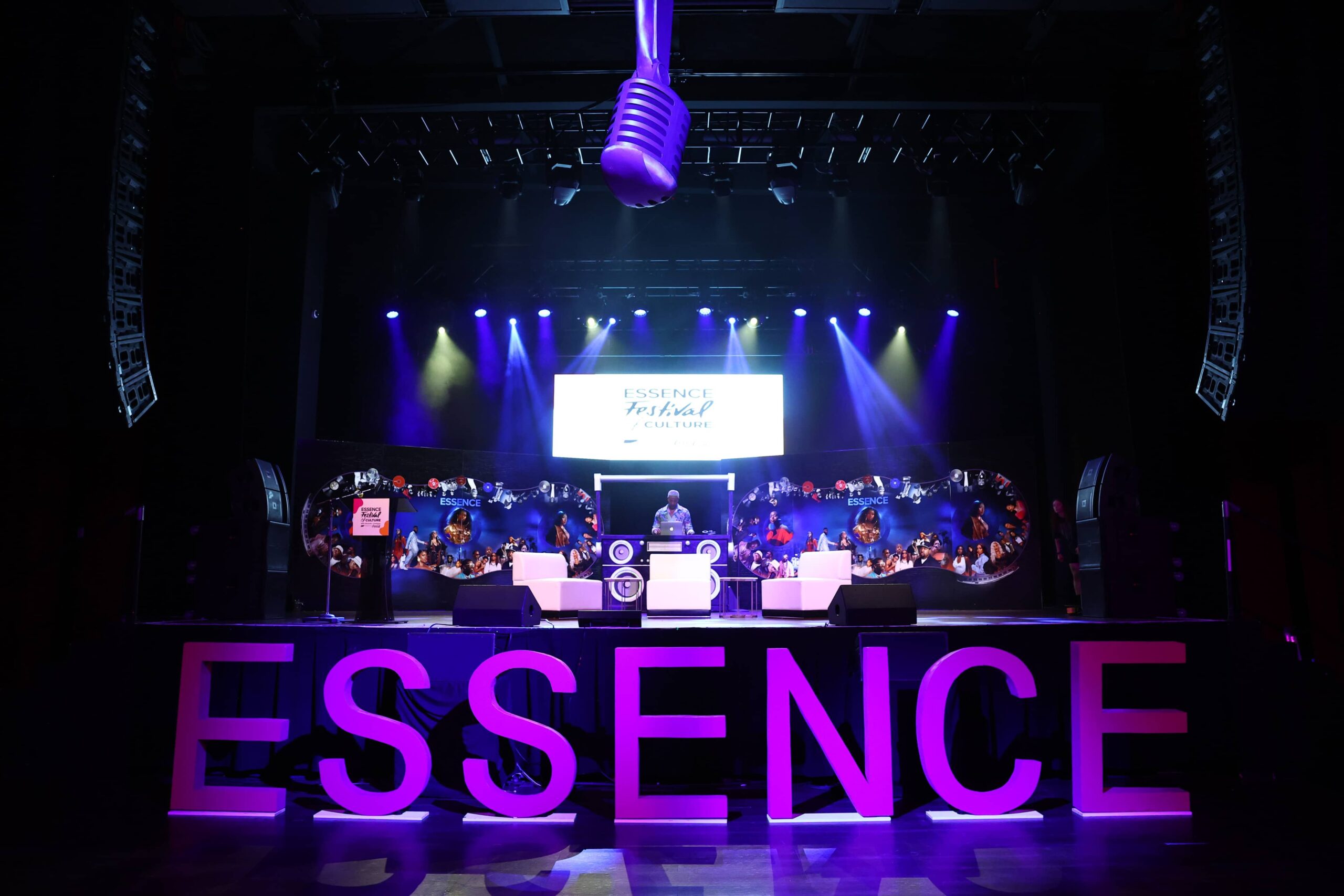 A stage with large purple ESSENCE letters, a DJ, bright spotlights, and a screen displaying “ESSENCE Festival” under a hanging microphone prop.