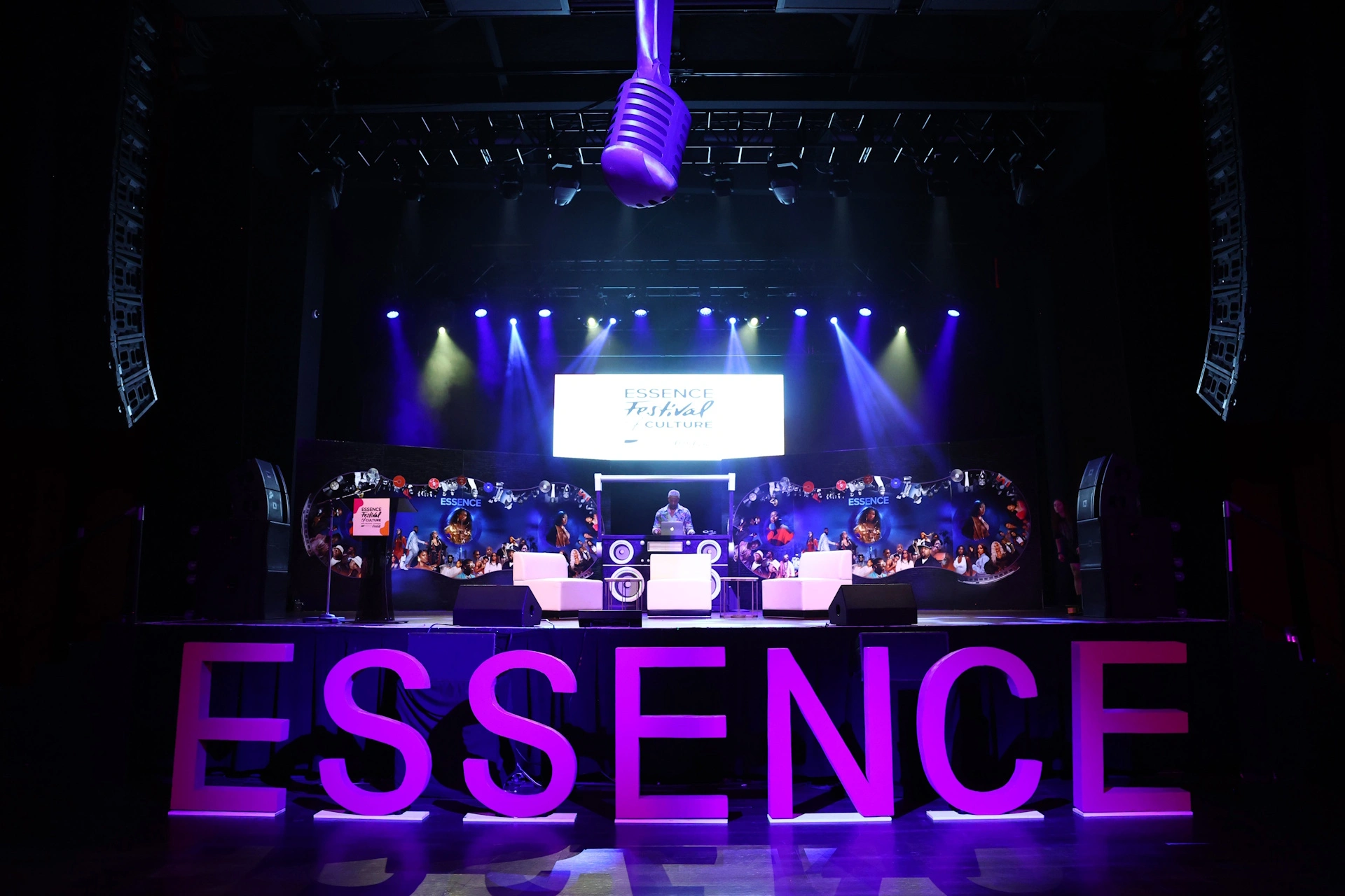 A stage setup with large purple "ESSENCE" letters, a DJ booth, and a backdrop displaying "ESSENCE Festival." Purple lighting and a large microphone prop hang overhead.