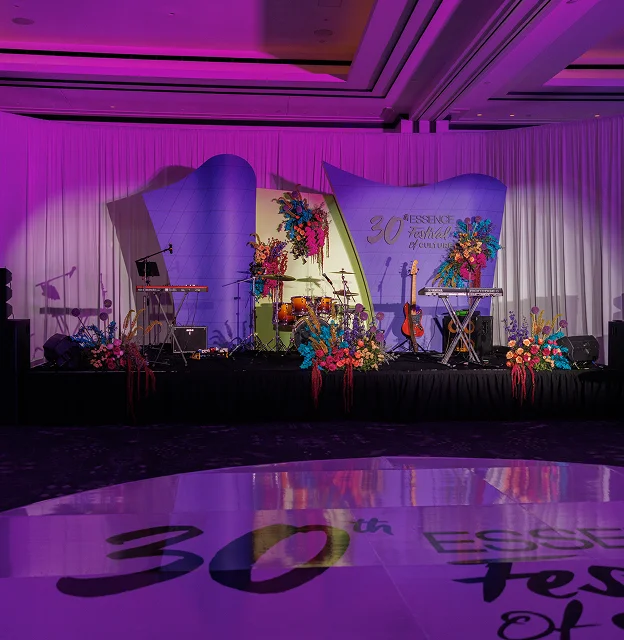 A stage with musical instruments and colorful floral arrangements is set up for the 30th Essence Festival of Culture, with purple lighting and a decorated white backdrop.