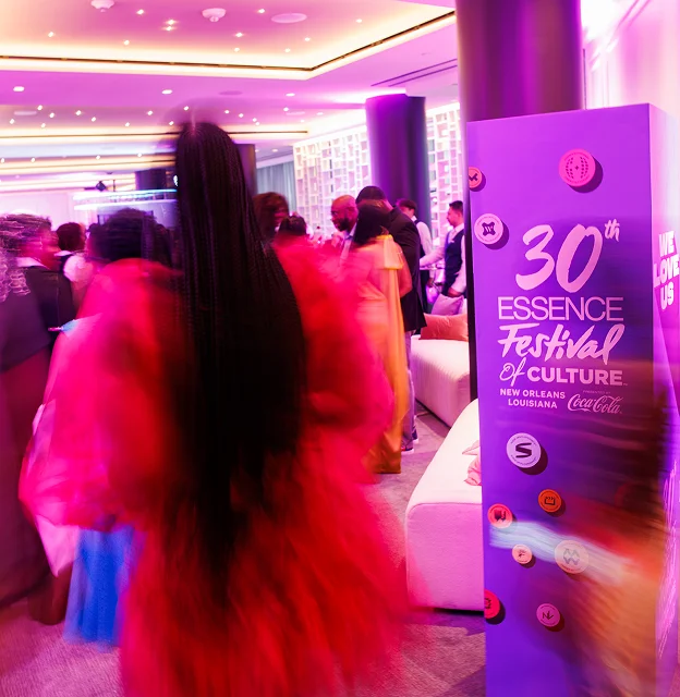 People attend an indoor event with pink lighting; a sign reads “30th Essence Festival of Culture, New Orleans, Louisiana, CocaCola.” A person in a red dress walks by.
