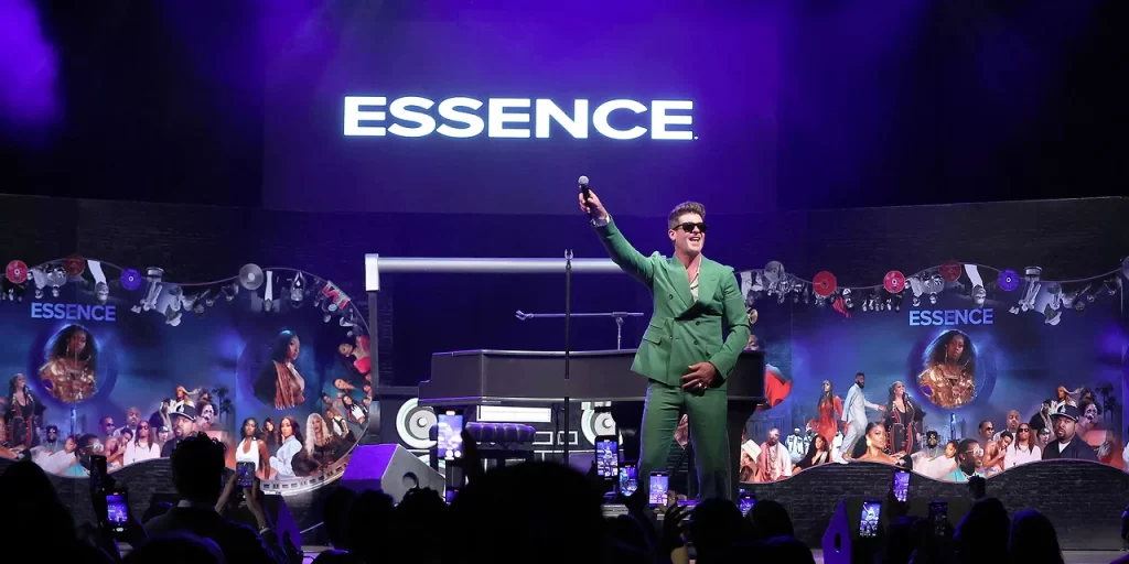 Performer in a green suit holds a microphone on stage at an Essence event, with audience members taking photos and event signage in the background.