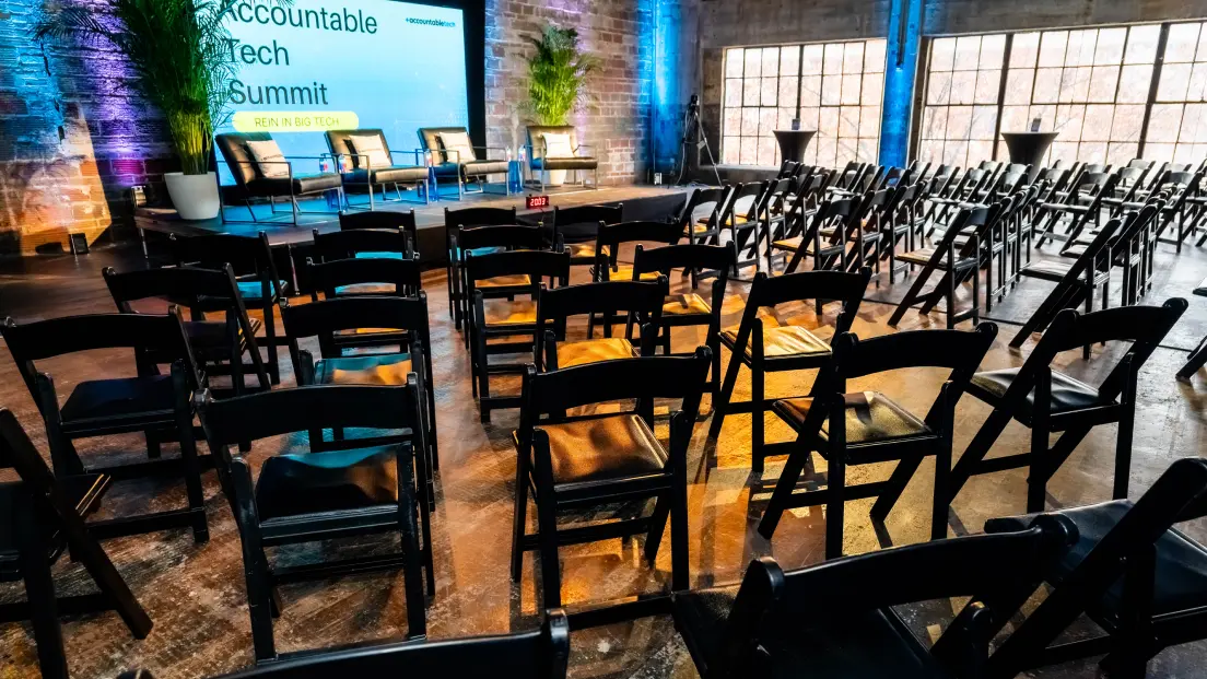 Rows of empty black chairs face a stage with four chairs and a screen displaying "Accountable Tech Summit" in an industrial-style event space.