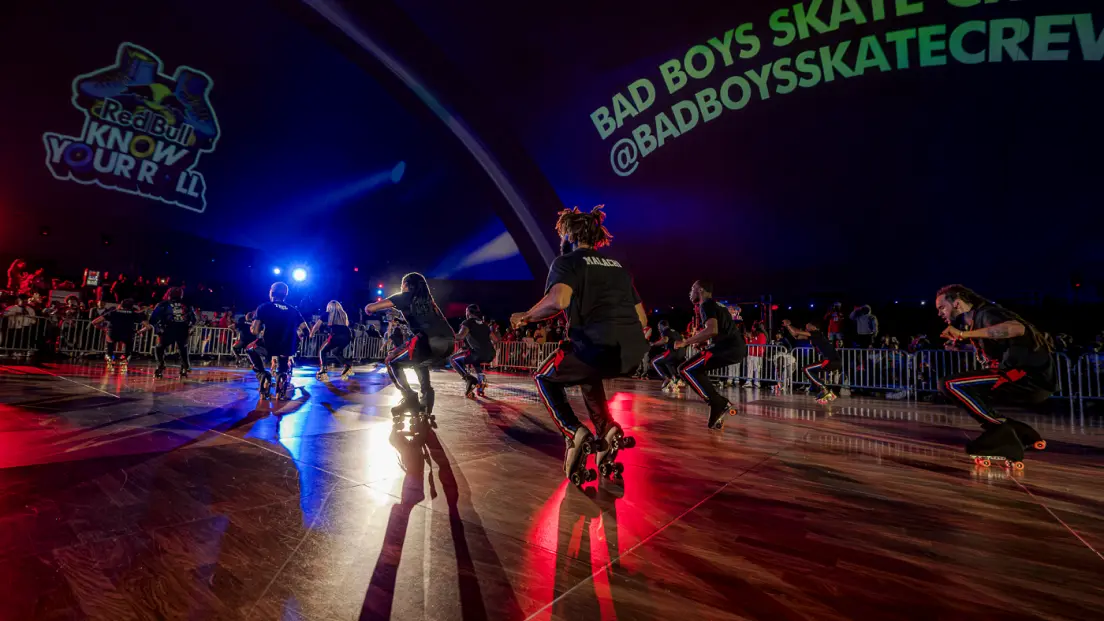 A group of people roller skating indoors under spotlights, with "Bad Boys Skate Crew" projected on the wall in the background.