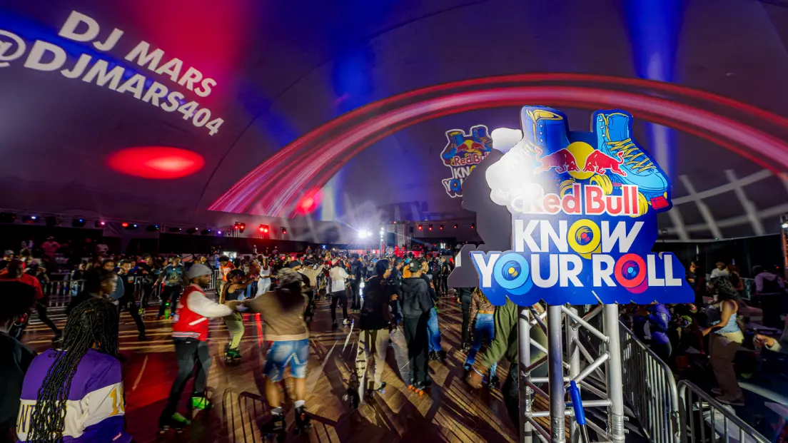 People roller skating at an indoor event with colorful lighting, “Red Bull Know Your Roll” sign in the foreground, and DJ Mars projected on the ceiling.