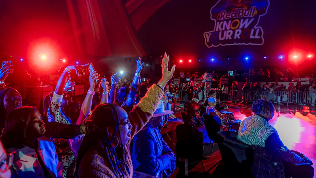 Audience members raise their hands and take photos at an indoor event with red and blue stage lights and a "Red Bull Know Your Roll" sign projected on the wall.
