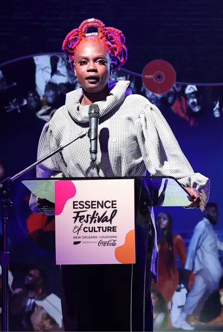 A person with bright red hair stands at a podium labeled "Essence Festival of Culture" in front of a dark backdrop with event photos.