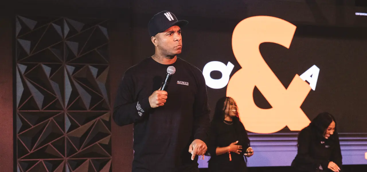 Man holding a microphone stands on stage during a Q&A event, with two people and a large “Q&A” sign in the background.