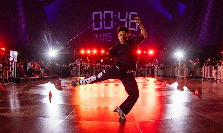 A person performs a mid-air dance move in a spotlight on a stage, with a digital timer reading 00:45 in the background and an audience watching behind barriers.