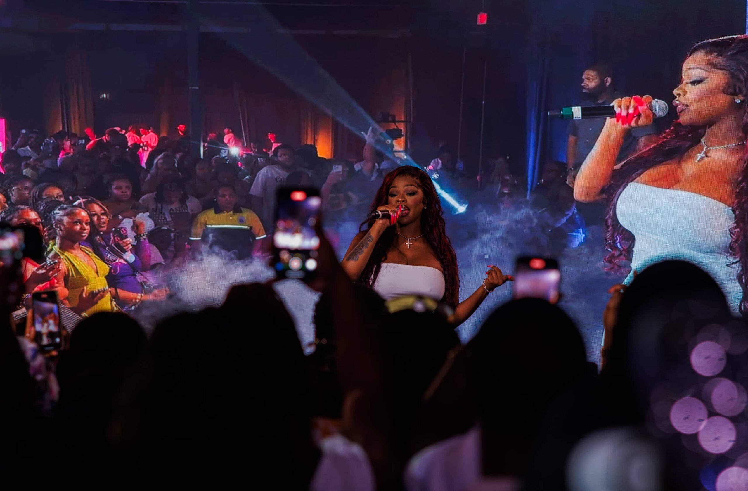 A singer performs on stage in front of a large crowd, with her image displayed on a big screen behind her. Audience members are filming with their phones.