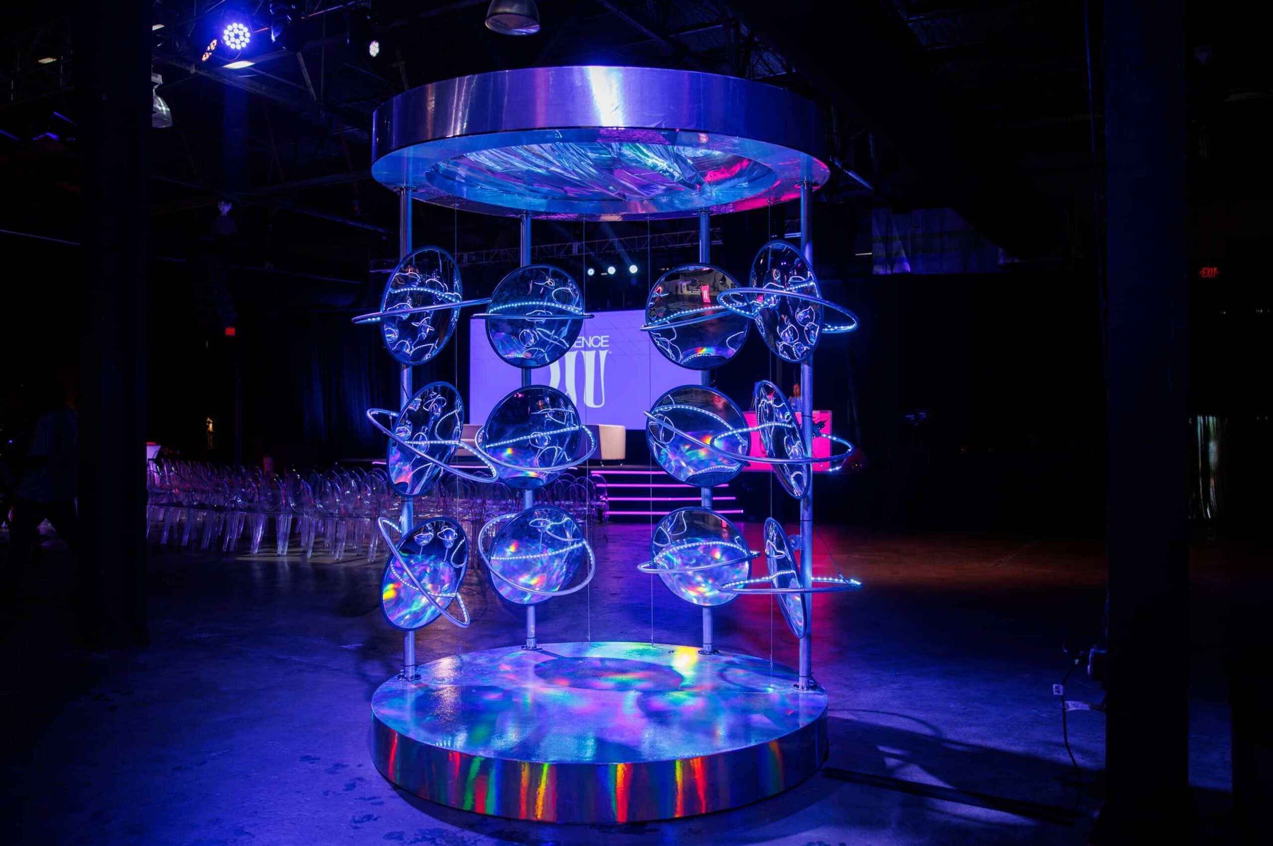 A large metallic art installation with circular elements and blue lighting stands in a dark event space; empty chairs and a stage are visible in the background.