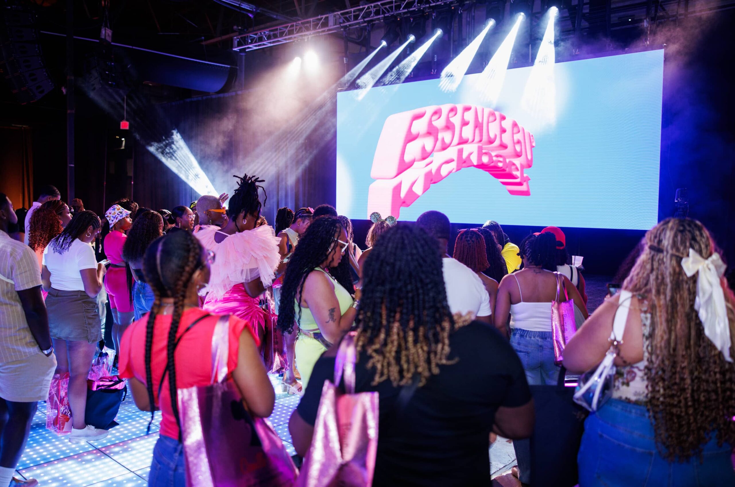 A crowd faces a stage with bright lights and a large screen displaying "Essence Kickback" at an indoor event.