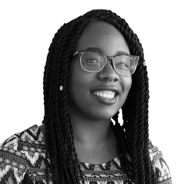 A person wearing glasses and patterned clothing smiles at the camera. They have long twist braids and a nose ring.