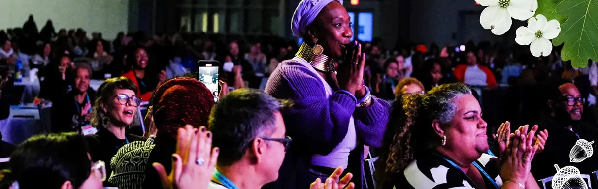 A diverse audience sits and applauds at an indoor event; a woman in purple stands and claps, with floral decorations in the image’s upper right corner.