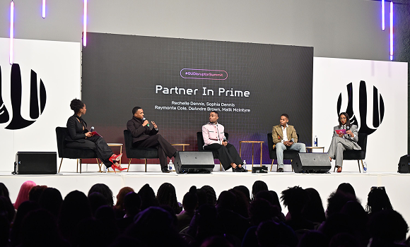 Five panelists sit on stage having a discussion at a live event called “Partner In Prime,” with an audience seated in front of them.