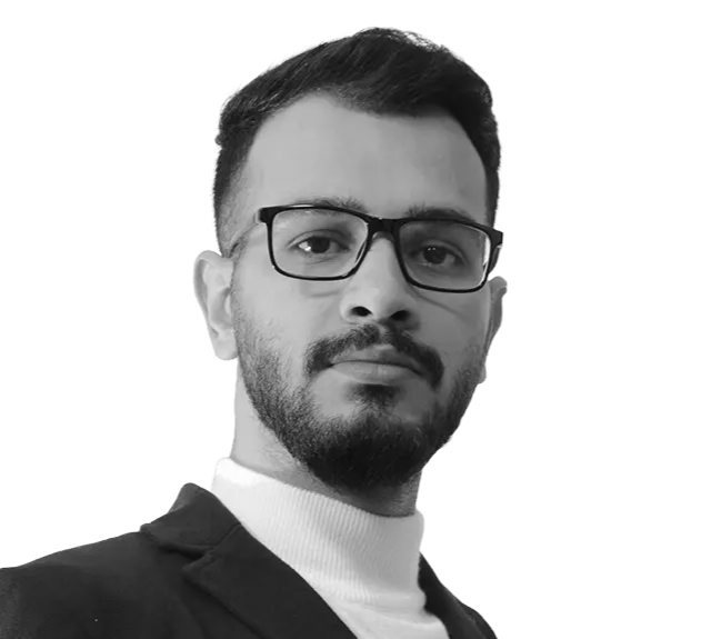 A man with short dark hair, glasses, and a beard wears a suit jacket and turtleneck, looking at the camera against a plain white background.
