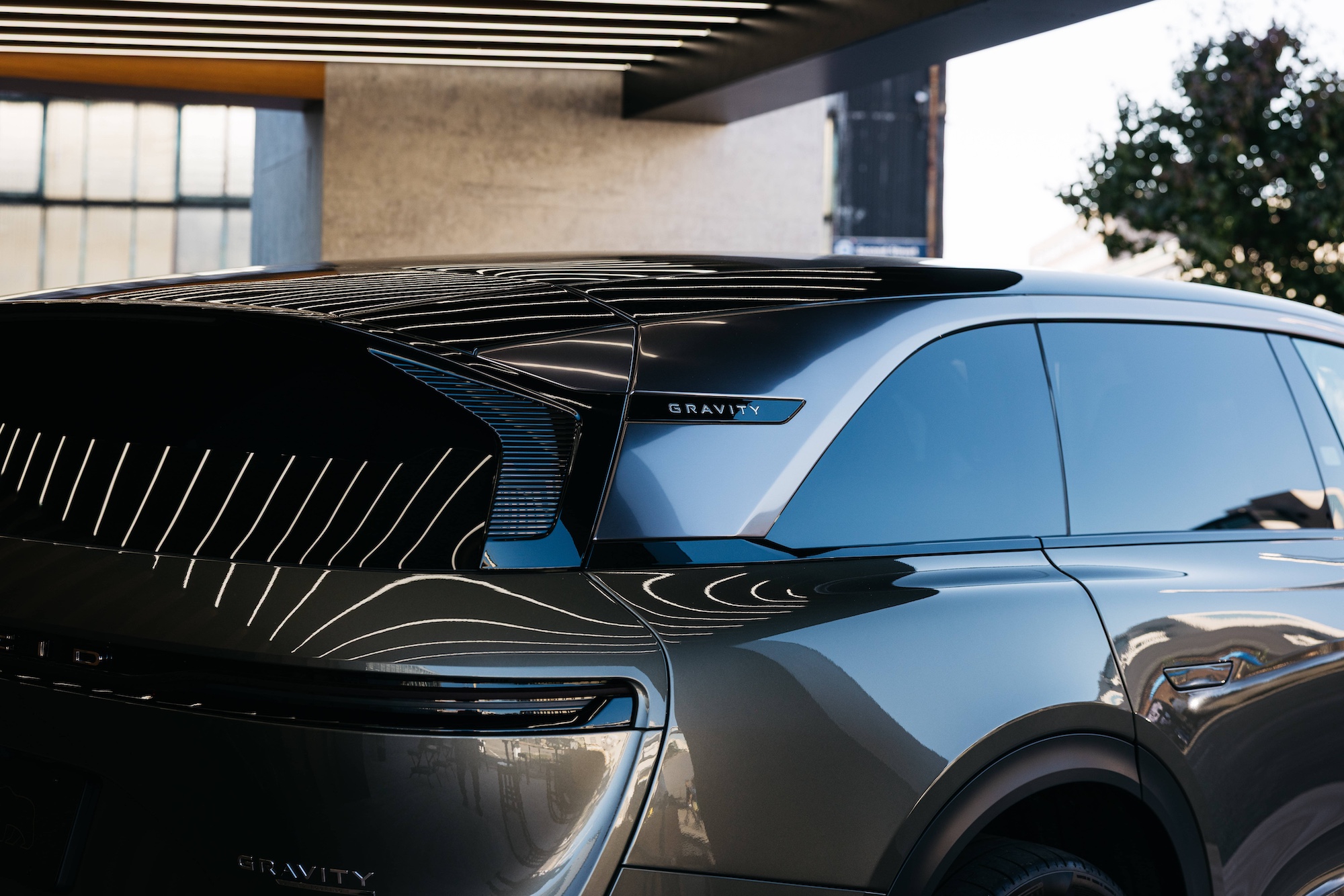Close-up of the rear side and window of a silver SUV with “Gravity” branding, parked under a structure with striped ceiling lights reflecting on the car’s surface.