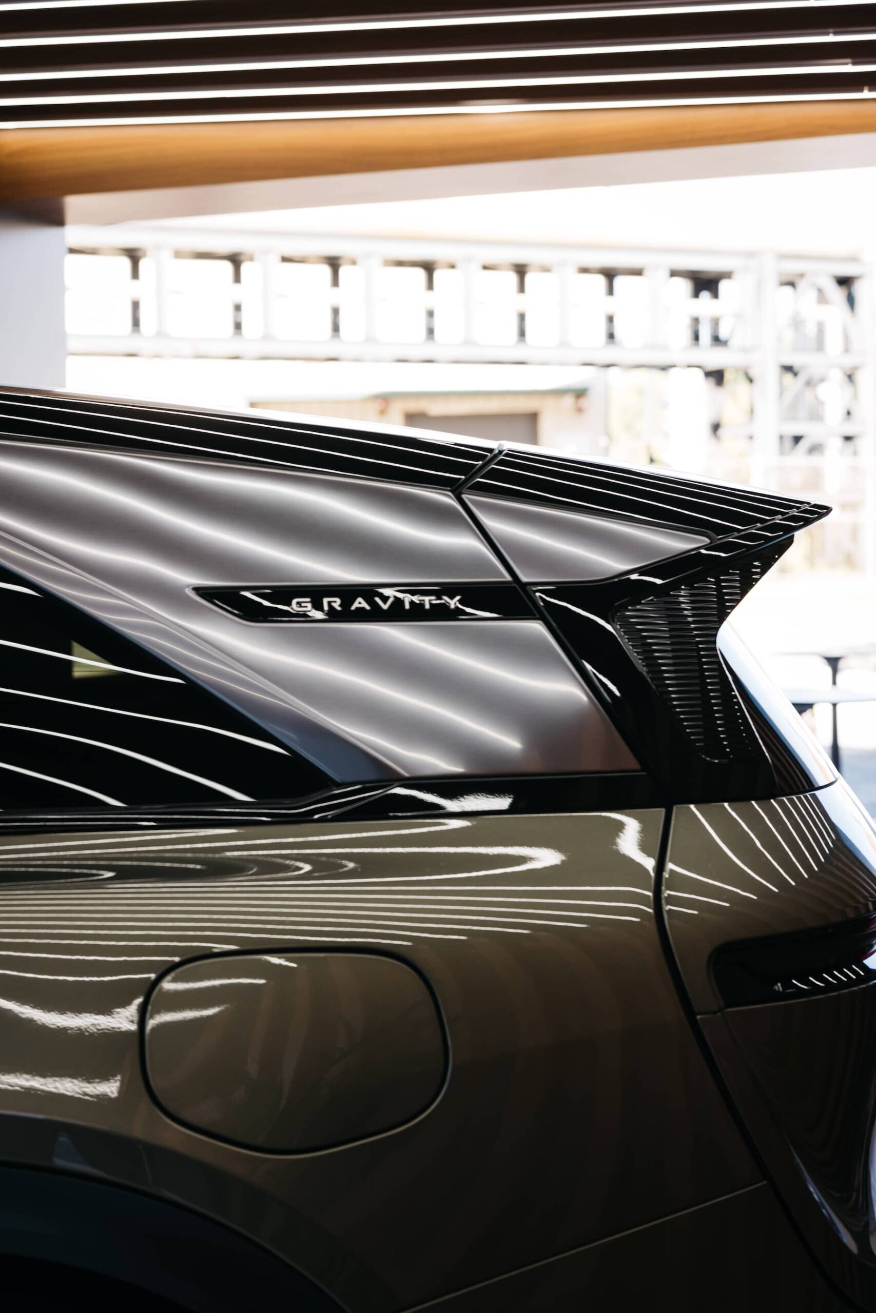 Close-up of the rear side and window of a car with the word "GRAVITY" on the trim, reflecting ceiling lights and a blurred building in the background.