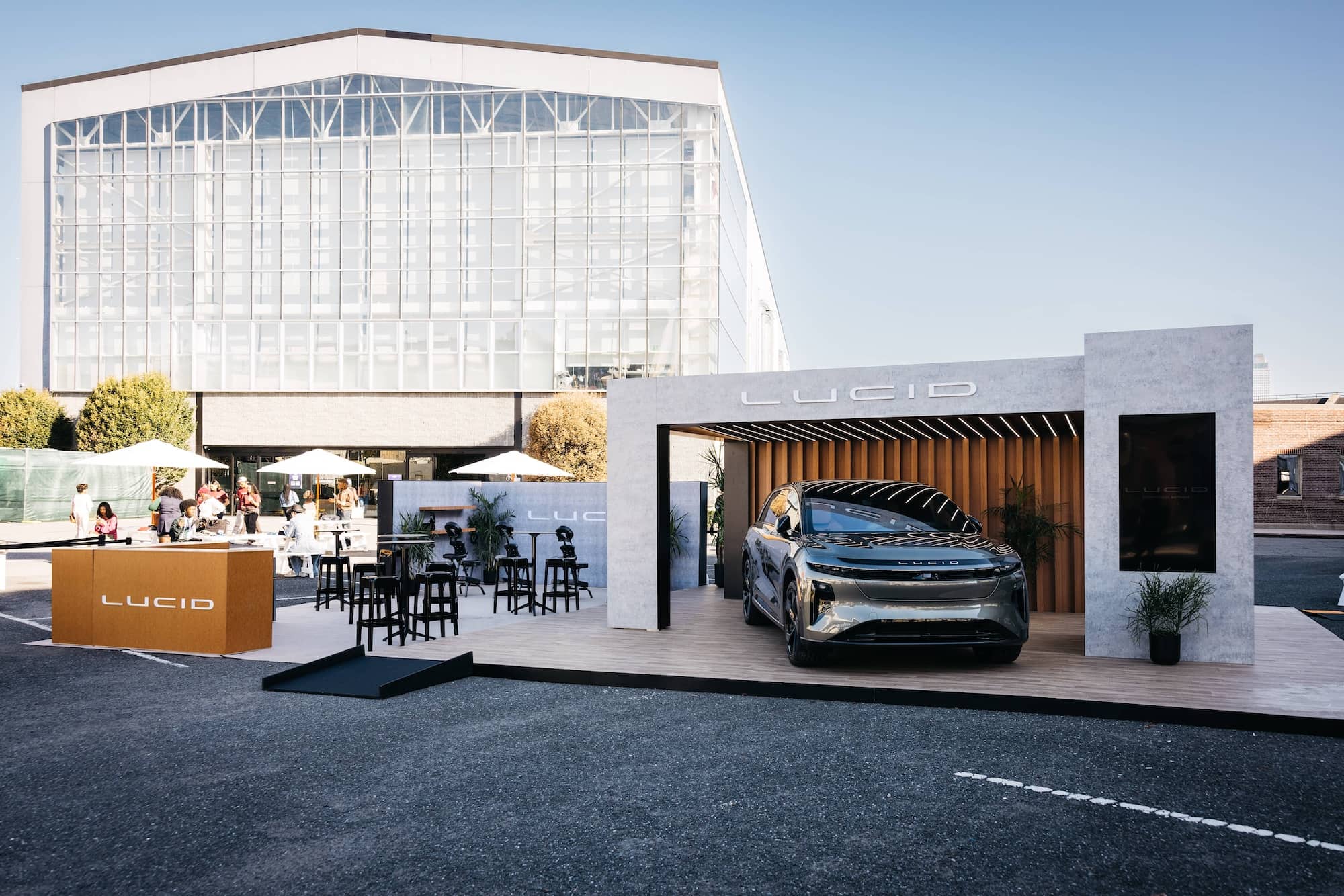 A Lucid electric car is displayed on a platform at an outdoor event, with tables, chairs, and people nearby. A modern building stands in the background.