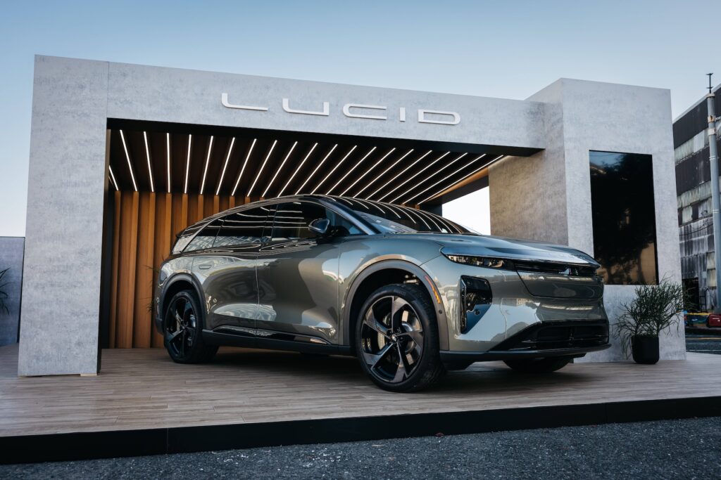 A metallic Lucid SUV is displayed on a wooden platform beneath a modern pavilion with a “LUCID” sign, outdoors under clear skies.