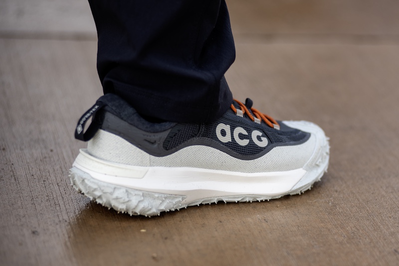 A person wearing black pants and a grey Nike ACG sneaker with orange laces stands on a brown surface.