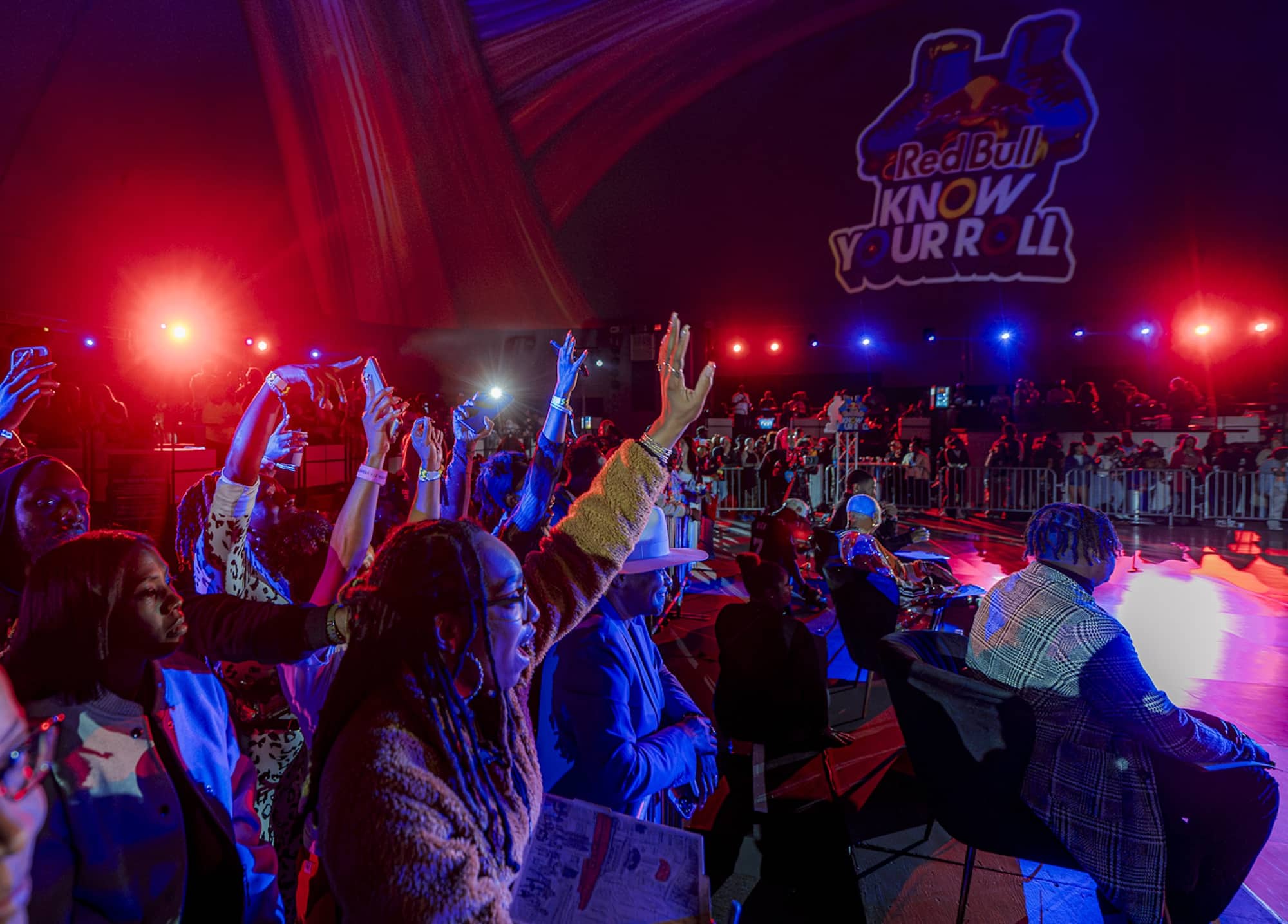 Audience members raise hands and take photos at a brightly lit Red Bull Know Your Roll event, with a large logo projected on the wall.