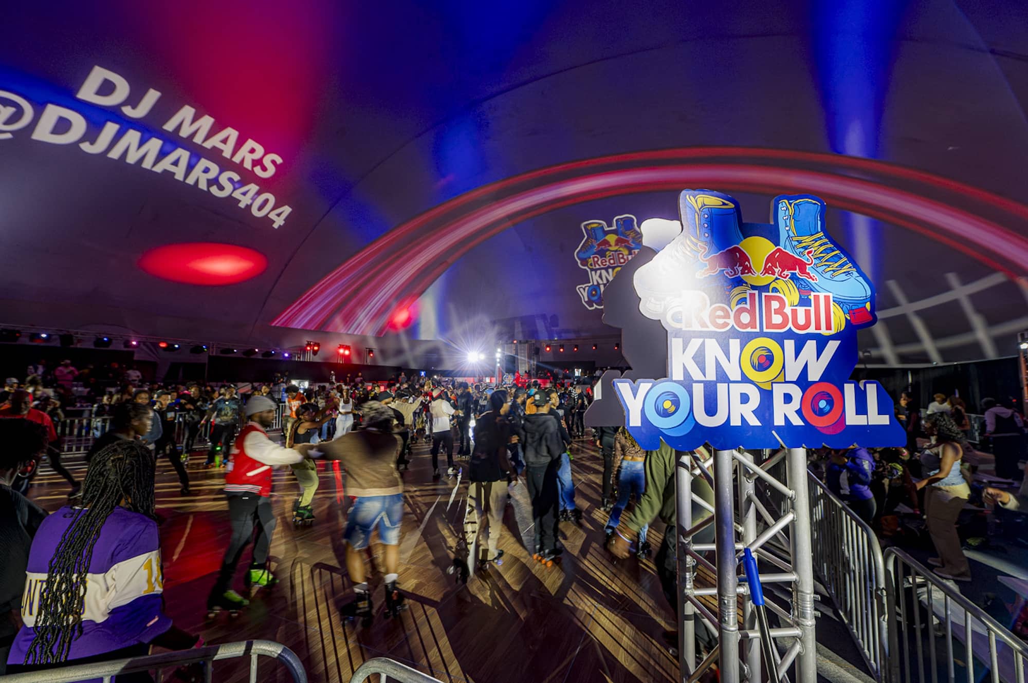 A crowd gathers at an indoor roller-skating event with a "Red Bull Know Your Roll" sign and DJ Mars' handle projected on the wall.