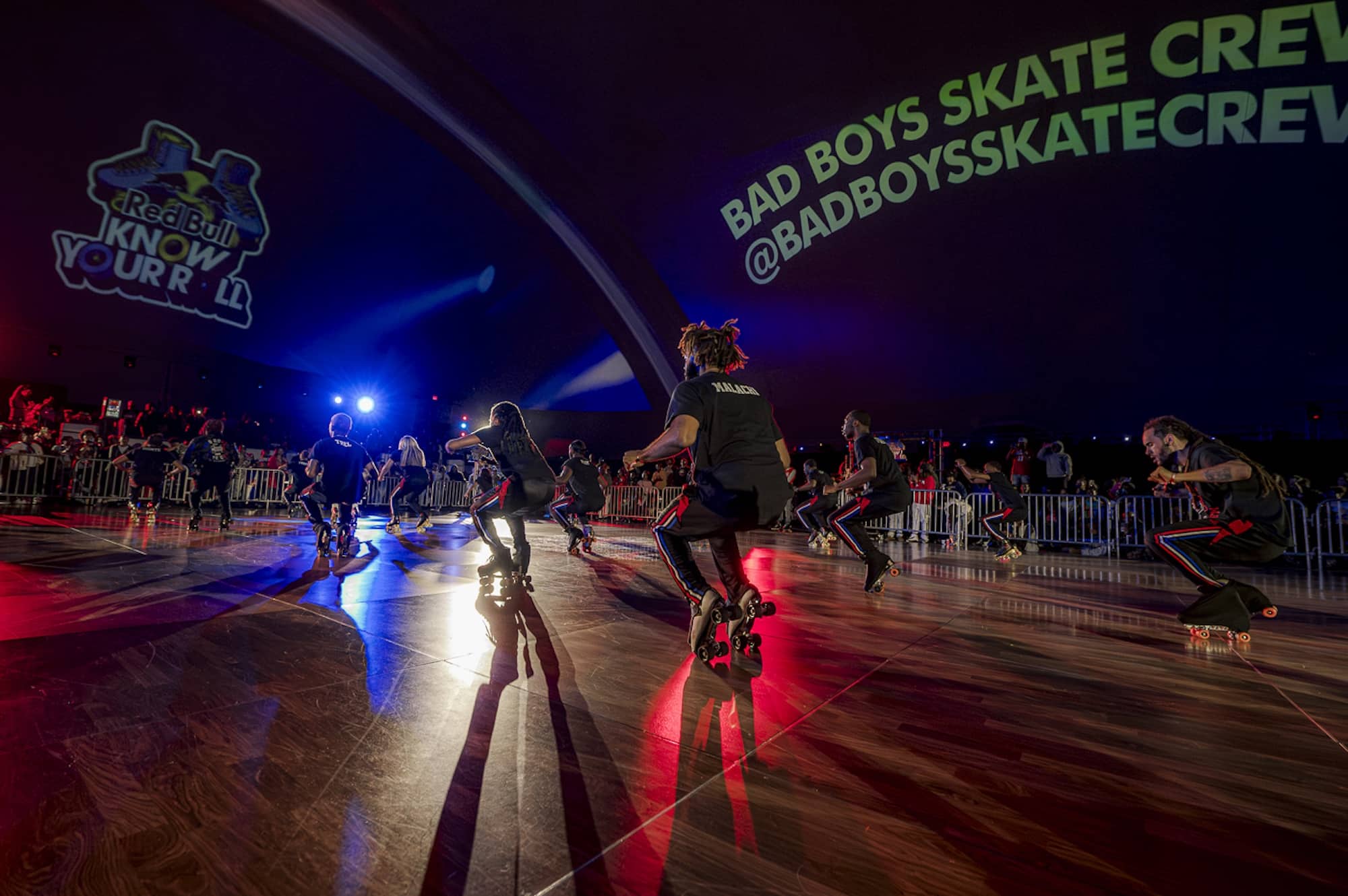 A roller skating group performs indoors on a lit stage with "BAD BOYS SKATE CREW @BADBOYSSKATECREW" and event branding projected on the wall.