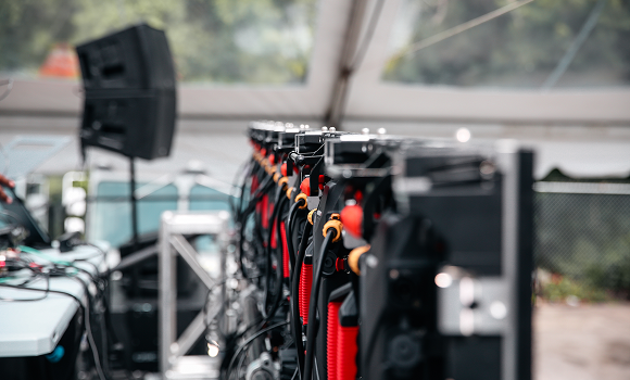 A row of professional audio speakers and equipment is set up under a tent outdoors, with cables and control panels visible.