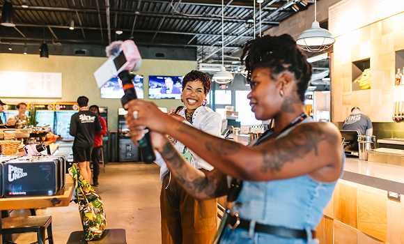 Two women take a selfie with a phone in a busy, modern restaurant. One woman smiles at the camera while the other holds the phone on a selfie stick in the foreground.